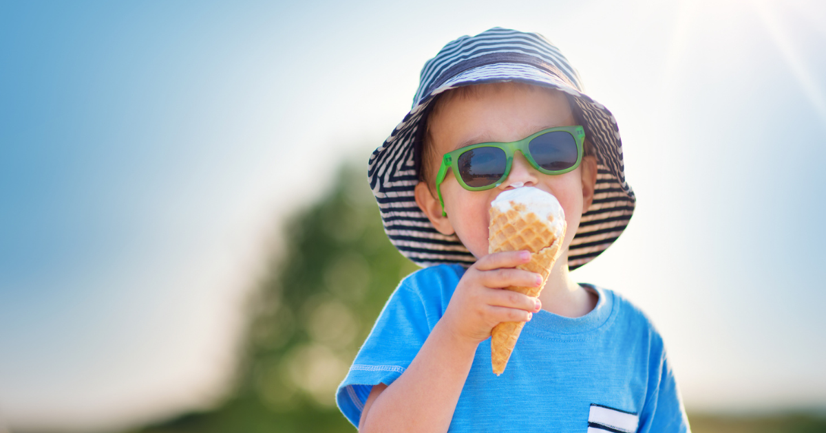 A little boy eating ice cream wearing a hat and sunglasses to protect him from the sun.