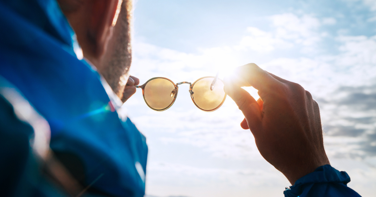 A man outside holding up his sunglasses to the sky.