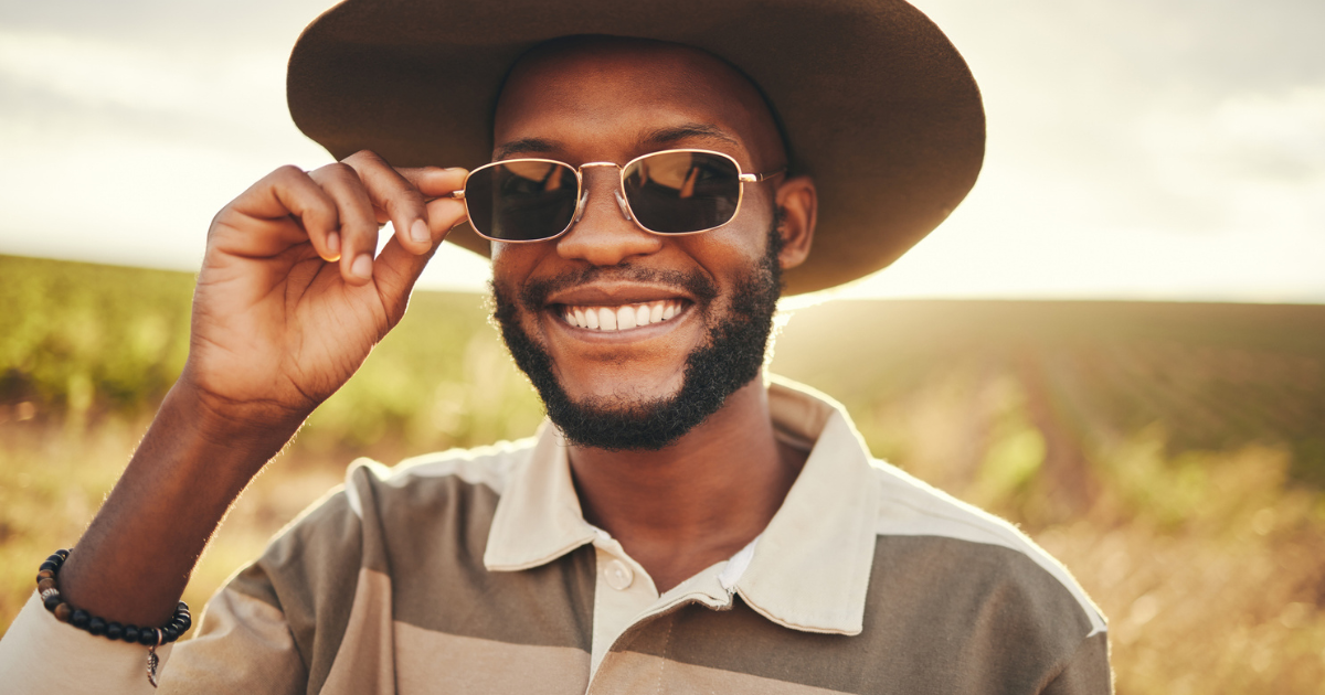 A black man smiling in the sun wearing a hat and sunglasses to protect against UV light damage.